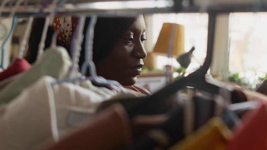 Selective focus close-up of attentive young African American woman browsing clothes on hangers in womenswear section at second hand outlet, examining dress and blouse, searching for stylish bargains