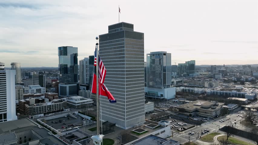 American flag and flag of Tennessee on flagpole in front of downtown skyline in Nashville city. Skyline with skyscrapers and Tennessee Tower with also blowing flag of United States on top of building.