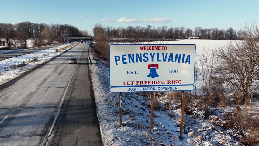 Aerial view of snowy highway at Pennsylvania state border, with the “Welcome to Pennsylvania” sign, winter traffic, frozen fields and clear blue sky. Maryland border in USA.
