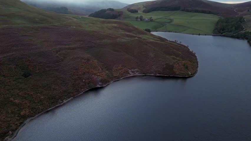 Aerial view of a calm lake surrounded by rolling green hills during evening light. Peaceful rural landscape with clear sky and natural textures, captured in autumn from above.