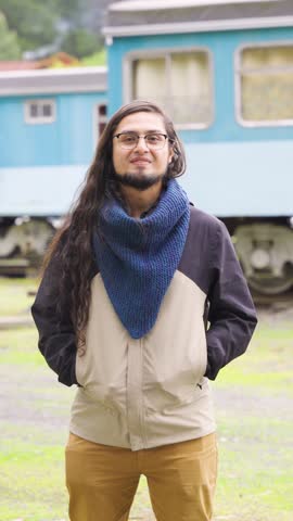 Speaking young man with long wavy hair and glasses, wearing a blue hand-knitted neck warmer and a casual jacket in front of a blue vintage train car.