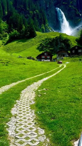 A stunning vertical shot of a powerful waterfall cascading down a forested mountain. In the foreground, traditional wooden houses sit on a vibrant green meadow with a stone-paved path leading toward t