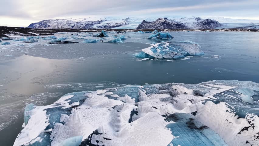 Aerial drone pullback over drifting icebergs in an Icelandic glacier lagoon, revealing snow-covered mountains on the horizon, calm glacial water and soft sunrise reflections