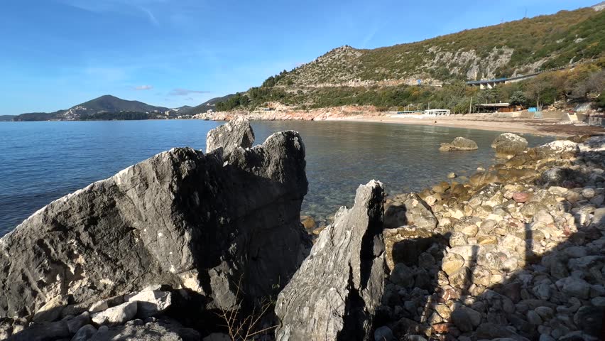 Rocky shoreline with large stones and pebbles along a calm bay, with clear water,
distant hills, and a quiet coastal landscape under a blue sky.