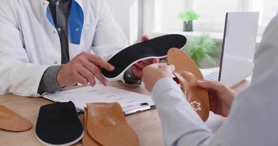 Orthopedist doctor shows orthopedic insoles to patient in medical clinic. They discuss custom orthotics and arch support to relieve foot pain. Concept medicine, foot healthcare and comfort.