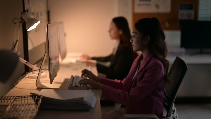 Two focused female employees typing on keyboards and working late in a dimly lit office environment