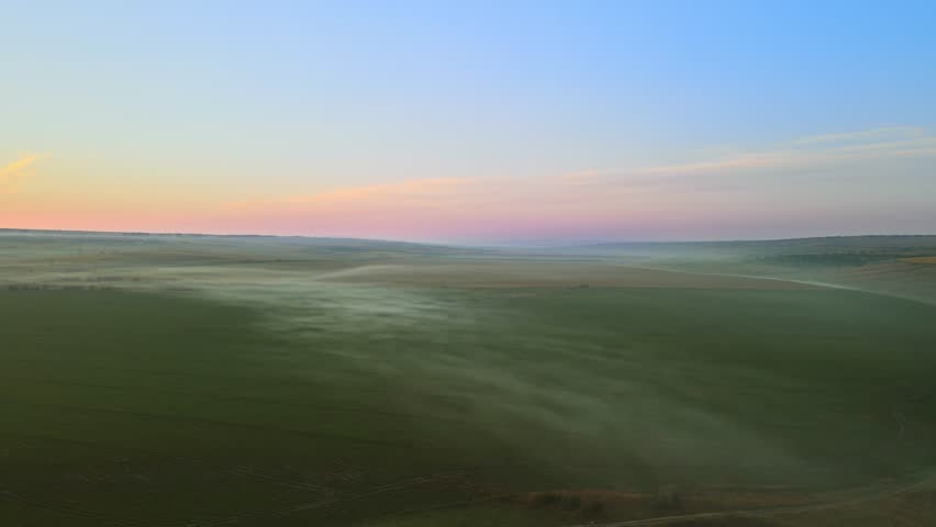 Aerial view of scenic sunrise over agricultural land with fields and a lake, revealing light fog moving above green vegetation