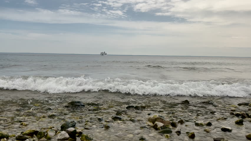 A peaceful, low-angle shot of crystal clear Lake Huron water washing over smooth stones on the coast of Mackinac Island, Michigan. A large passenger ship is visible on the distant horizon under a bright summer sky.