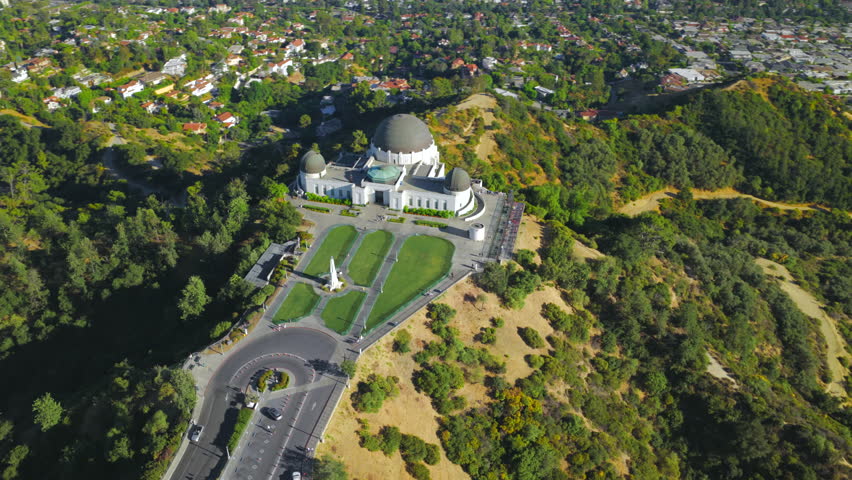 Aerial drone view of Griffith Observatory on Mount Hollywood with green lawns, surrounding hills and expansive Los Angeles California cityscape under clear daylight sky.