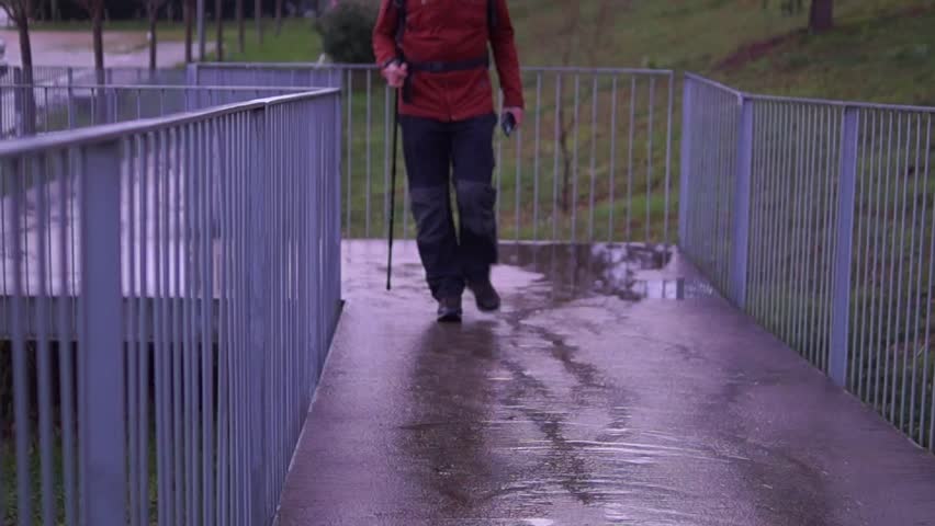 A man in hiking attire walks towards the camera on a wet concrete path, holding a trekking pole and checking his phone in a rainy outdoor setting.