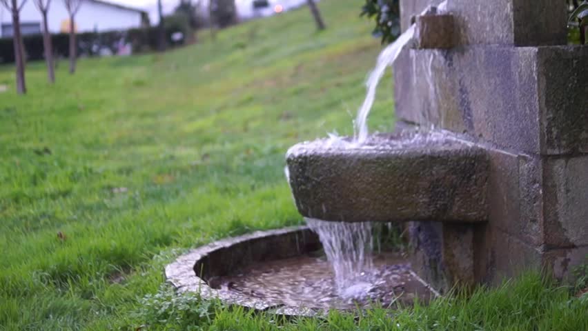 Close-up of a solo traveler wearing a red jacket washing his hands with fresh water from a rustic stone fountain in a park.