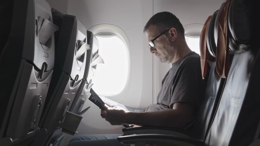 A man sits by the window on an airplane and reads a magazine. He is focused on the pages while the plane is in the air. The sunlight brightens the cabin.