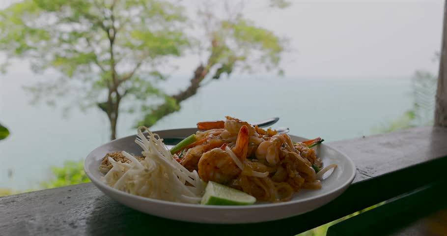 Pad Thai with shrimp resting on wooden balcony tropical ocean. Authentic Thai cuisine and luxury travel experience.