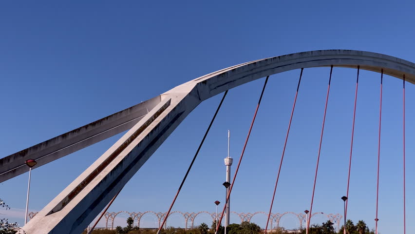 Close view of contemporary arched bridge in Seville spanning the calm river below View of the Guadalquivir River with trees in foreground and Alamillo Bridge rising against bright blue sky