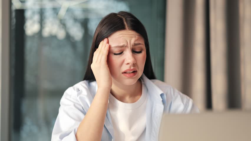 Young woman experiencing severe headache, showing signs of stress and discomfort. Manifests exhaustion and pain, struggling with wellness.