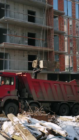 Red KamAZ truck being loaded with large concrete block by crane. Construction site operation showcasing heavy machinery in action.