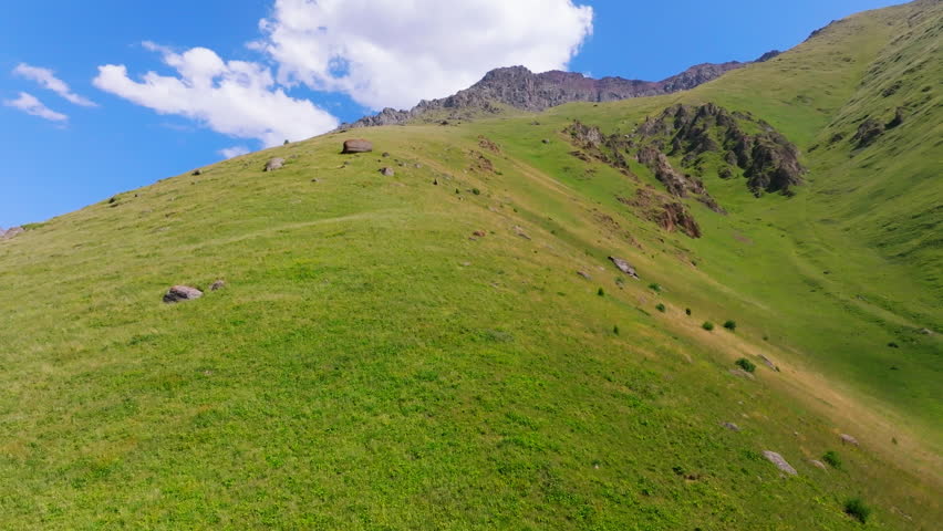 View from a drone ascending uphill while flying parallel to a steep grassy mountainside at constant low altitude above the terrain. Summer sunny day.
