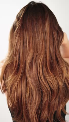 VERTICAL VIDEO: Brunette woman shakes her long hair in a beauty salon. View from behind of a young woman with wavy brown hair. Natural texture. Hair salon advertisement.