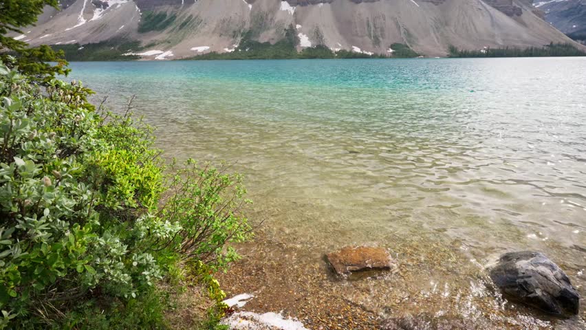 The camera panning up to reveal the stunning, glacier-fed turquoise Bow Lake in Alberta, Canada, in Banff National Park with waves gently lapping against shore in front of huge shear mountains