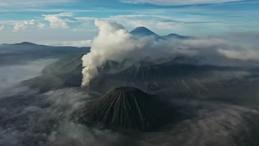 Aerial view of volcanic landscape Bromo Tengger Semeru National Park, Indonesia. Smoke rises from active volcano among cloud formations and distant mountain range under bright sky.