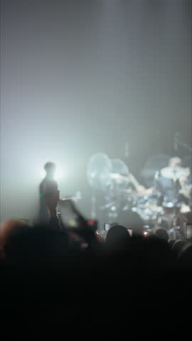 Silhouettes of a crowd at a live music concert. Fans hold up phones to record the show. The stage is lit with dramatic red lights, creating a vibrant, energetic atmosphere. Vertical video.