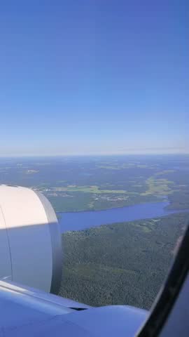 Aerial view from an airplane window flying over a beautiful landscape of lakes and green forests under a clear blue sky.