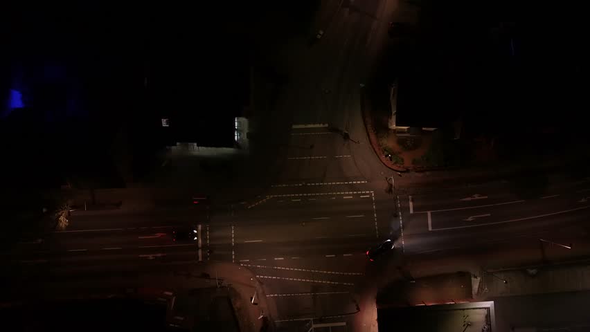 Black car turning right on junction of American town at night. Aerial top down shot. warm glowing lantern and streetlamps in quiet city night. Tracking shot. Bright lighting headlights of vehicle.