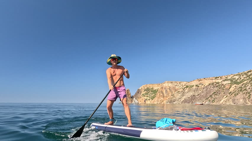 Paddleboarding man ocean, paddling on sup board with scenic cliff backdrop under clear sky