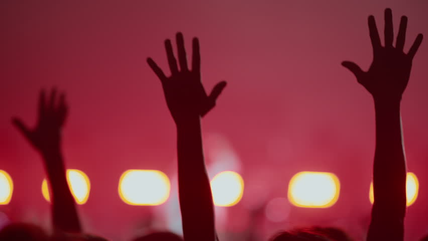 Silhouettes of a crowd with hands raised in the air at a live music concert or festival. Red stage lights and smoke create an atmospheric and energetic scene of celebration and worship.