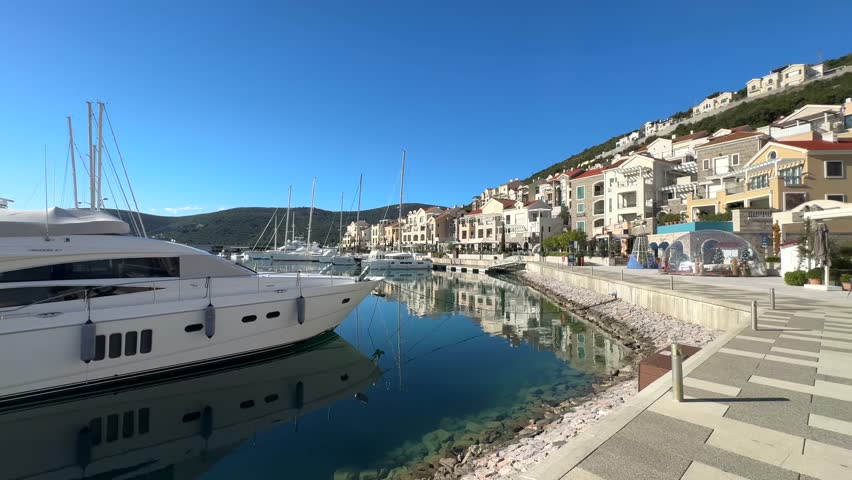 Modern marina with moored yachts along a waterfront promenade, calm water reflecting boats and residential buildings under a clear blue sky.