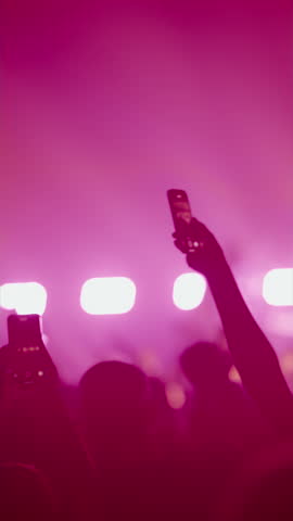 Vertical shot of a crowd at a live music concert. Fans hold smartphones up, recording the show. Silhouettes are visible against bright, flashing stage lights and atmospheric smoke.