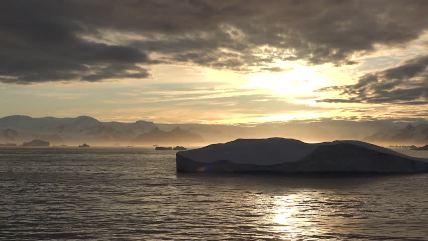 Icebergs move slowly in a blue fjord in Greenland. The sun shines brightly as snow-covered mountains surround the calm water. Nature shows its power in this icy landscape.