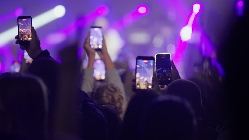 Footage of a crowd at a live music concert. Fans hold up smartphones to record the show, then raise their hands to cheer and dance. Silhouettes against bright purple and blue stage lights.