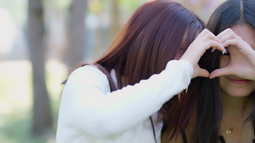 A happy, beautiful gay couple are smiling and making a heart shape with their hands in the park.