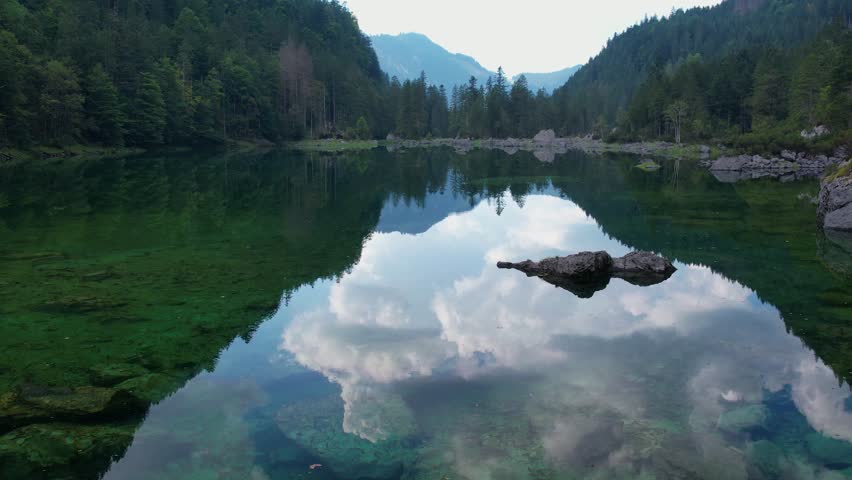 Explore a stunning aerial view of a tranquil lake surrounded by lush trees and mountains. The reflection of clouds creates a picturesque scene in nature.