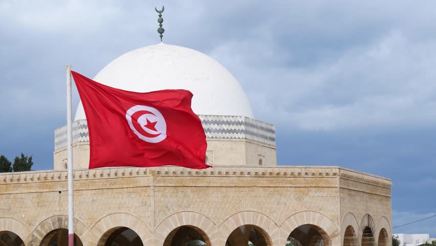 Tunisian flag waving in front of the Bourguiba Mausoleum in Monastir, Tunisia, featuring a white dome and arched stone architecture under a cloudy sky, symbolizing national identity and heritage
