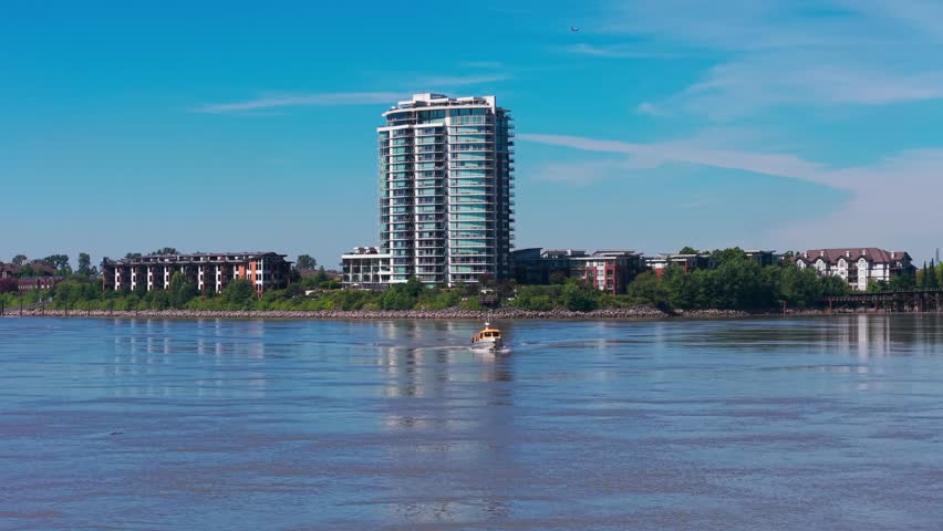 Drone view of New Westminster waterfront with a boat crossing the Fraser River, showcasing skyline, calm water and clear blue sky in British Columbia.