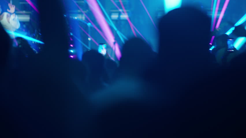 Soft-focus video of a crowd's silhouettes dancing and raising hands at a concert. Colorful blue, pink, and purple stage lights flash in the background, creating an energetic and atmospheric party