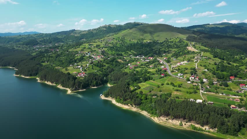 Cinematic backward drone shot over Lake Bicaz, Romania, revealing lush green hills, forested shoreline and a peaceful rural village—ideal for travel, nature, tourism and lifestyle visuals.