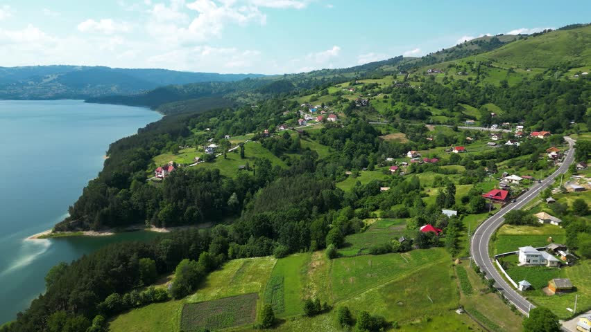 Aerial drone footage of Lake Bicaz region, Romania, showcasing green hills, scattered rural houses, winding roads and calm blue waters in a scenic mountain landscape. Drone going forward.