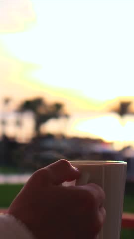 Close up of a female hand holding a white mug of hot coffee or tea with steam rising, enjoying a peaceful morning sunrise from a balcony with a beautiful, soft focus background