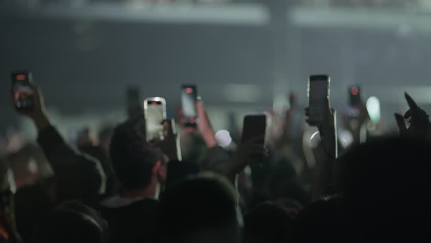 Video sequence of a crowd at a live concert filming the stage with their smartphones. The lighting changes dramatically from bright pink and red to dark, then to vibrant purple and pink.