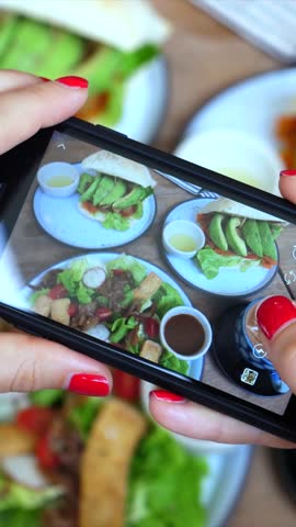 Female food blogger with red nails holding a smartphone and taking pictures of healthy vegan lunch in a cafe. Influencer creating social media content of tasty avocado toast and fresh salad
