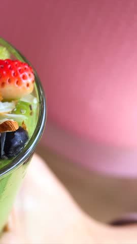 Close up of a vibrant green smoothie in a glass, topped with fresh strawberries, blueberries, kiwi, chia seeds, and sliced almonds, with a black straw being placed inside