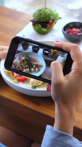 Food blogger taking a photo of pasta and a green burger with a mobile phone camera for her social media. Trendy culinary vlogging and content creation at a restaurant with healthy gourmet meal