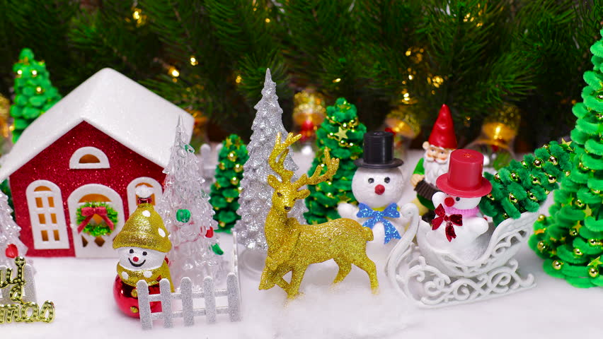 Near a red house surrounded by a snowman and Christmas trees and fenced with a fence with letters in front there is one yellow deer and snowmen with Christmas trees