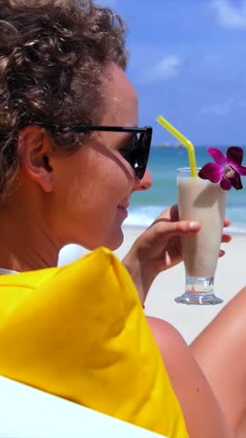 Attractive young woman with curly hair and sunglasses relaxing on a lounge chair by the sea, smiling and sipping a fresh fruit smoothie from a glass decorated with a purple orchid flower