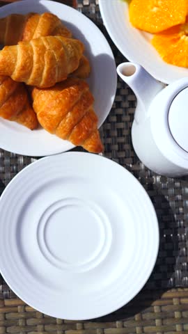 Overhead view of a person taking a bite out of toast with strawberry jam during a sunny morning breakfast, with croissants, fresh fruit, and a teapot on the wicker table