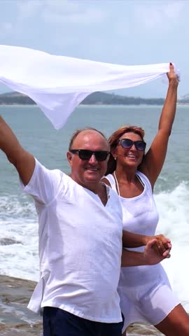 Joyful retired couple in white clothes standing on a rocky seashore, holding hands and a scarf blowing in the wind, celebrating their life together on a sunny day during a tropical vacation
