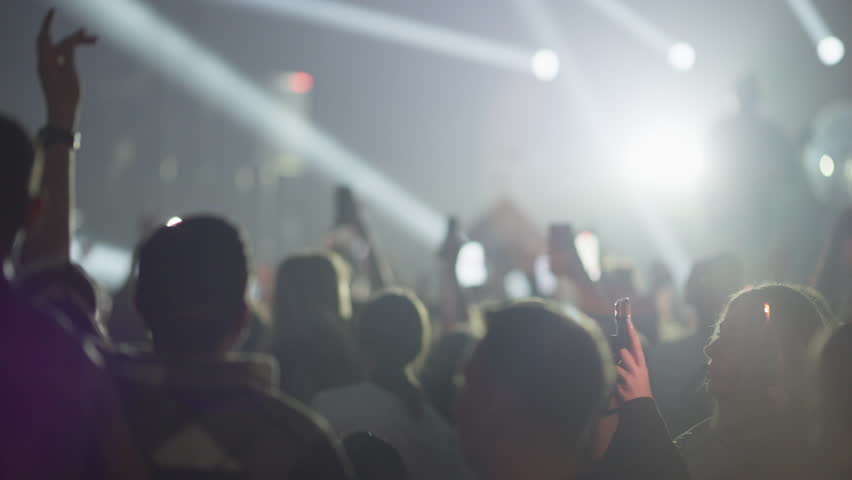 Video from within a crowd at a live music concert. Starts with white spotlights, transitions to vibrant purple stage lights as fans cheer and record on phones, and ends with a fade to black.
